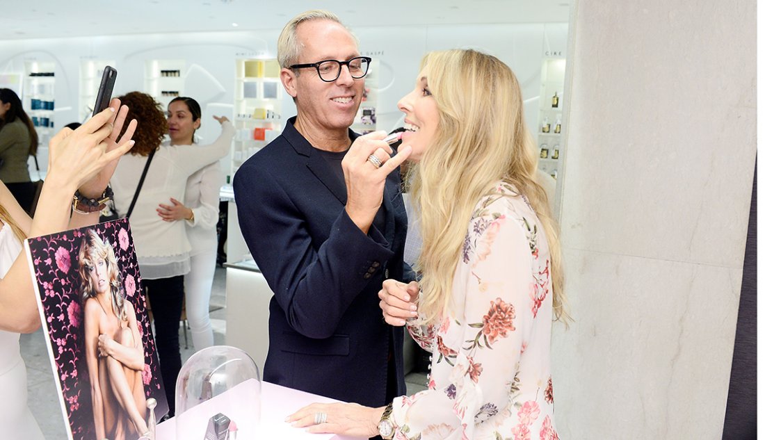 A Tim Quinn applies lipstick to Alana Stewart during a beauty event inside a bright, modern cosmetics store, with product displays and people in the background.