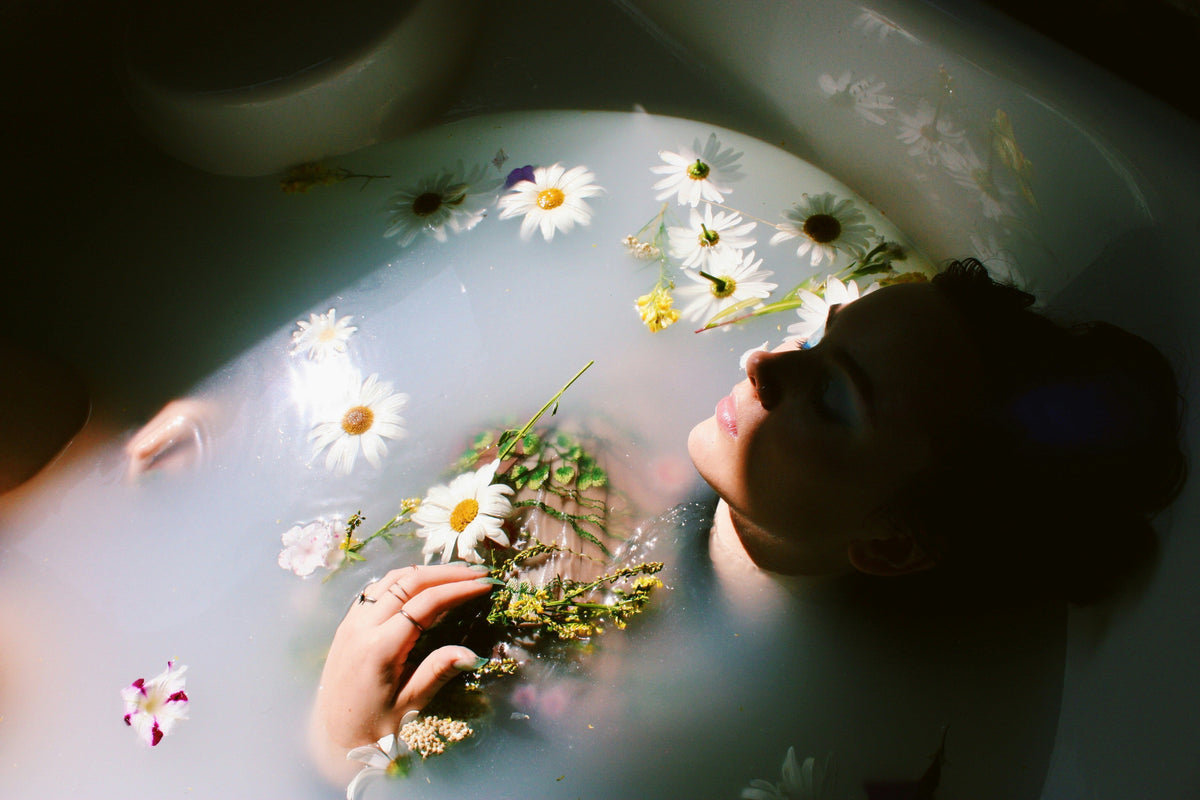 Woman seated in a botanical courtyard touching her neck and shoulder in warm sunlight.